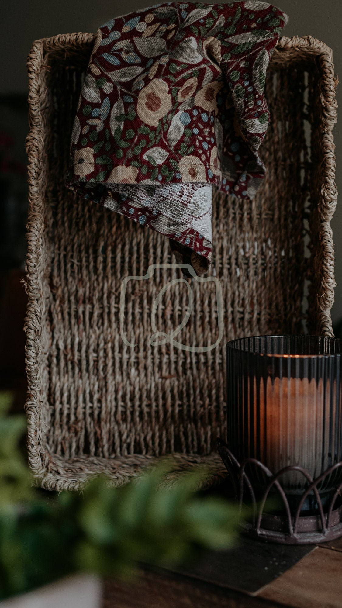 Woven basket with floral cloth and candle lantern beside plant, softly lit in cosy rustic scene