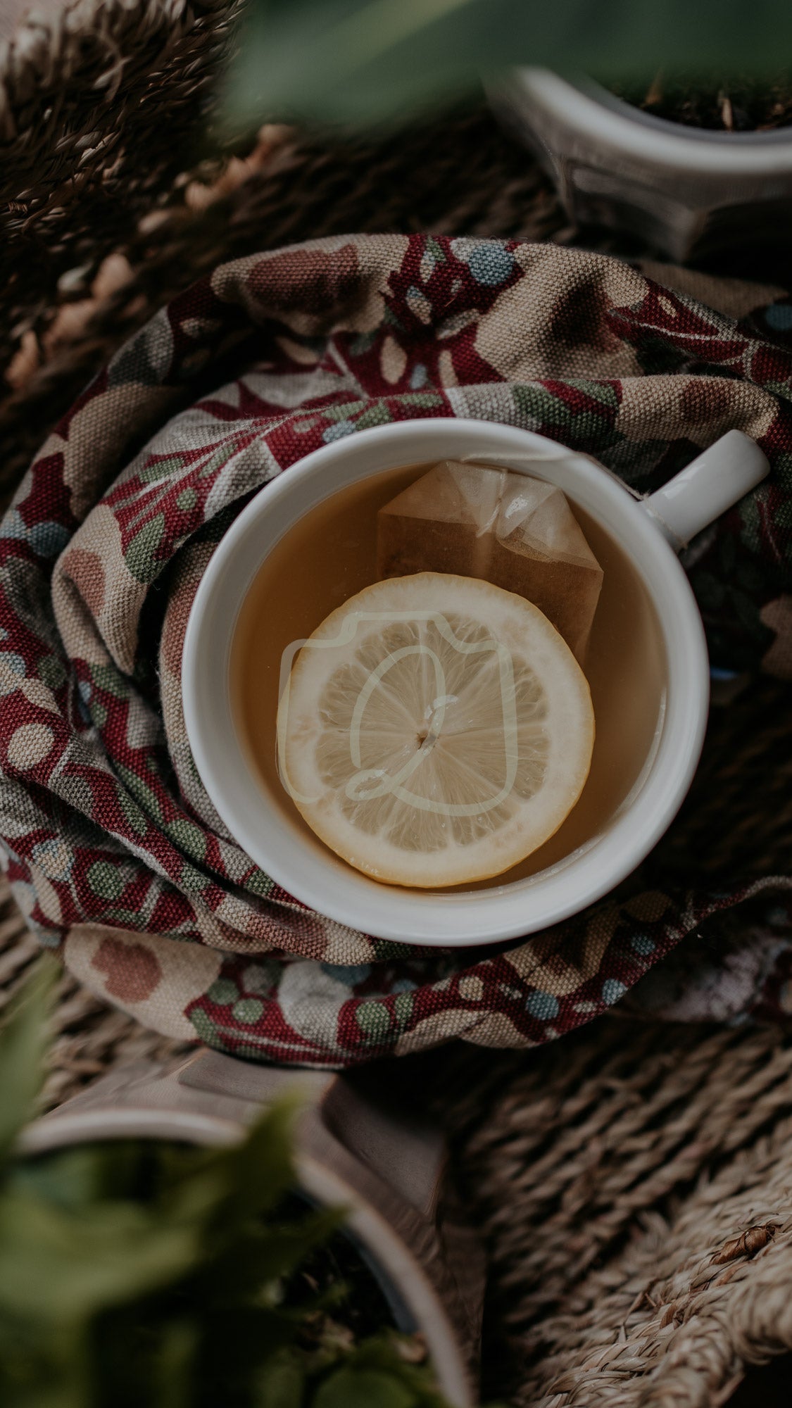 Cup of lemon tea with ice on patterned cloth and woven surface, surrounded by soft greenery