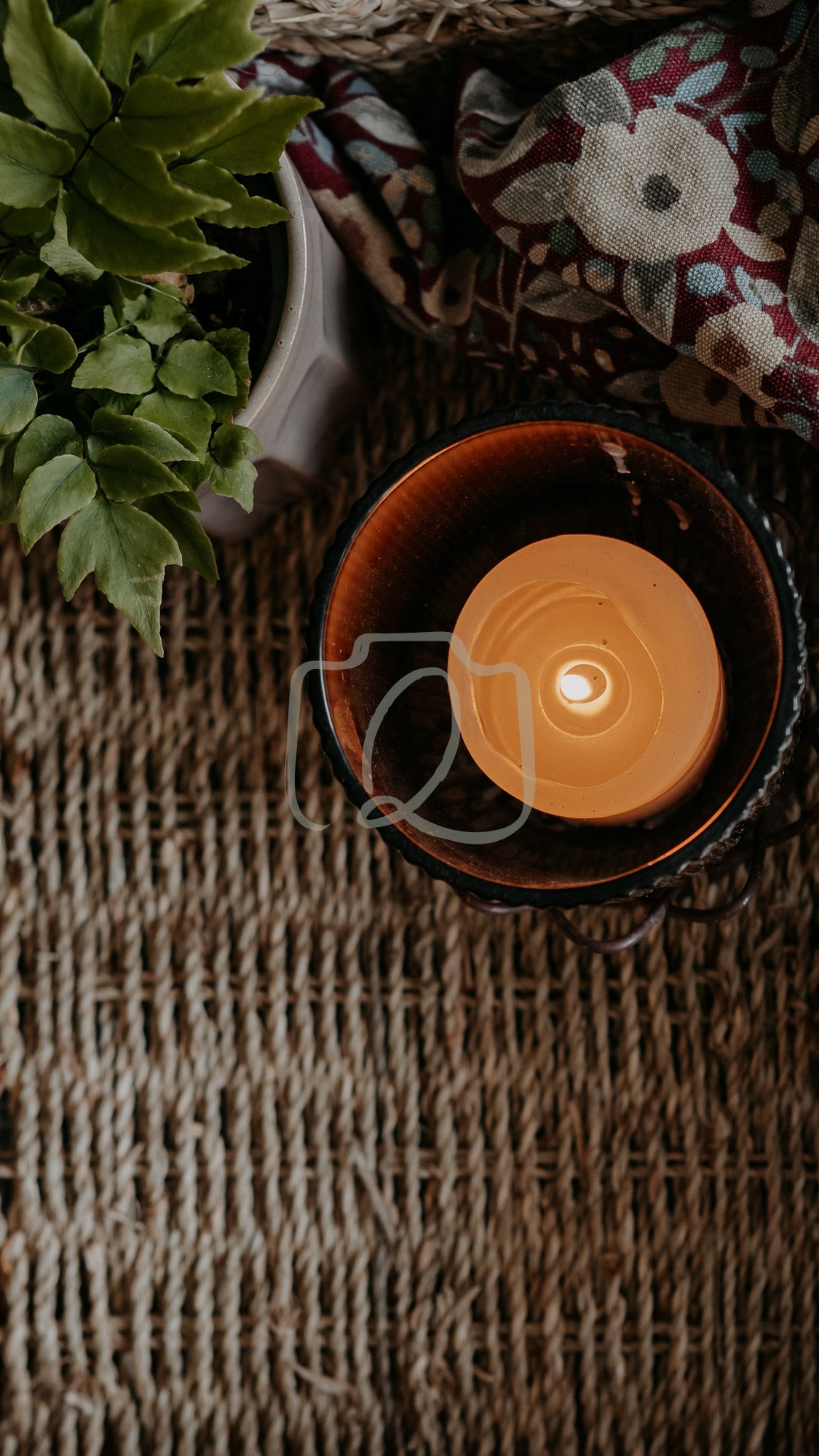 Lit candle in ceramic holder on woven surface with plant and patterned cloth in cozy scene